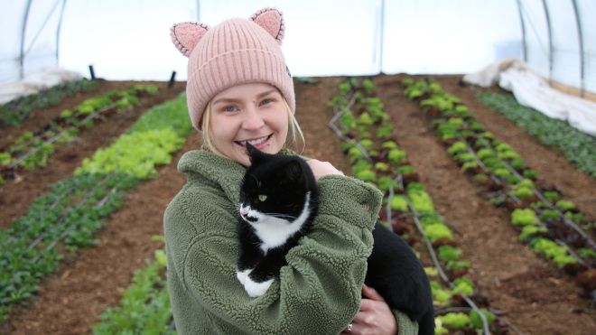 Sustainability Coordinator Kayla Tillapaugh stands in a greenhouse holding a cat and smiling